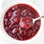 A white bowl filled with thick, chunky cranberry sauce, with a spoon scooping some of the sauce. The sauce is glossy and deep red in color.