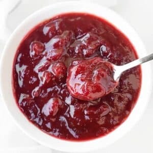 A white bowl filled with thick, chunky cranberry sauce, with a spoon scooping some of the sauce. The sauce is glossy and deep red in color.