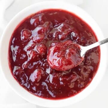 A white bowl filled with thick, chunky cranberry sauce, with a spoon scooping some of the sauce. The sauce is glossy and deep red in color.