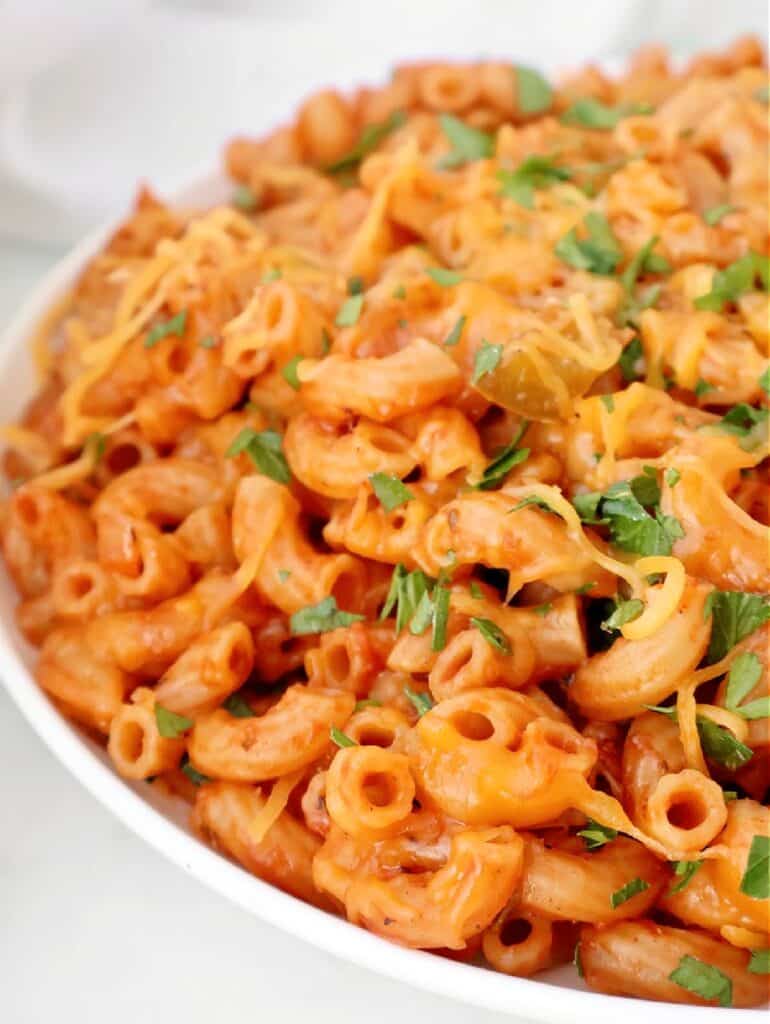A close-up of a bowl filled with creamy macaroni pasta in a tomato-based sauce, reminiscent of American goulash, topped with melted cheese and garnished with fresh chopped parsley.