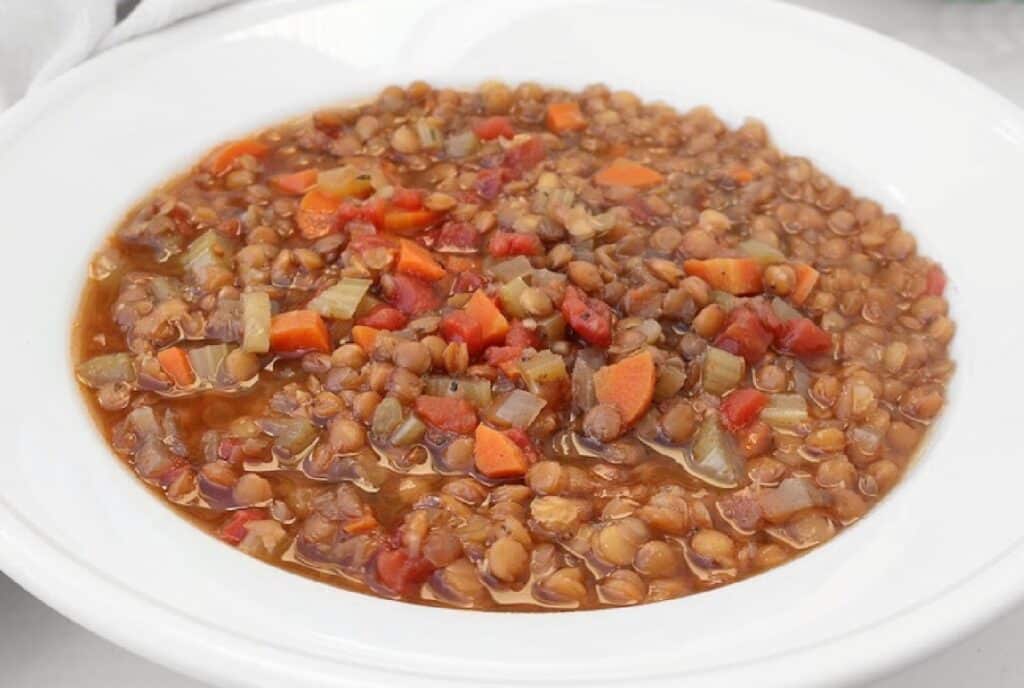 A white bowl filled with hearty lentil soup containing brown lentils, diced carrots, tomatoes, celery, and onions in a rich broth.