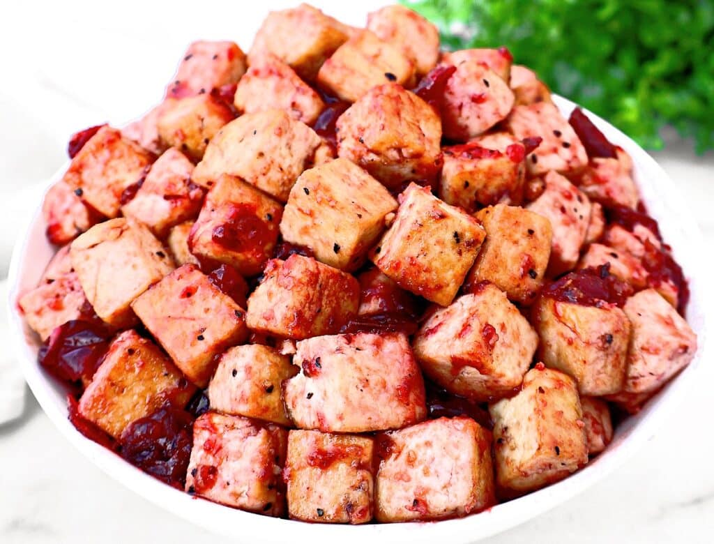 A bowl filled with cubed tofu coated in a reddish sauce, garnished with black pepper, with fresh green herbs blurred in the background.