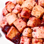 Close-up of golden-brown tofu cubes coated in a glossy red sauce, served in a white bowl. Some sauce pools around the tofu, giving a vibrant and appetizing appearance.