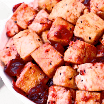 Close-up of golden-brown tofu cubes coated in a glossy red sauce, served in a white bowl. Some sauce pools around the tofu, giving a vibrant and appetizing appearance.