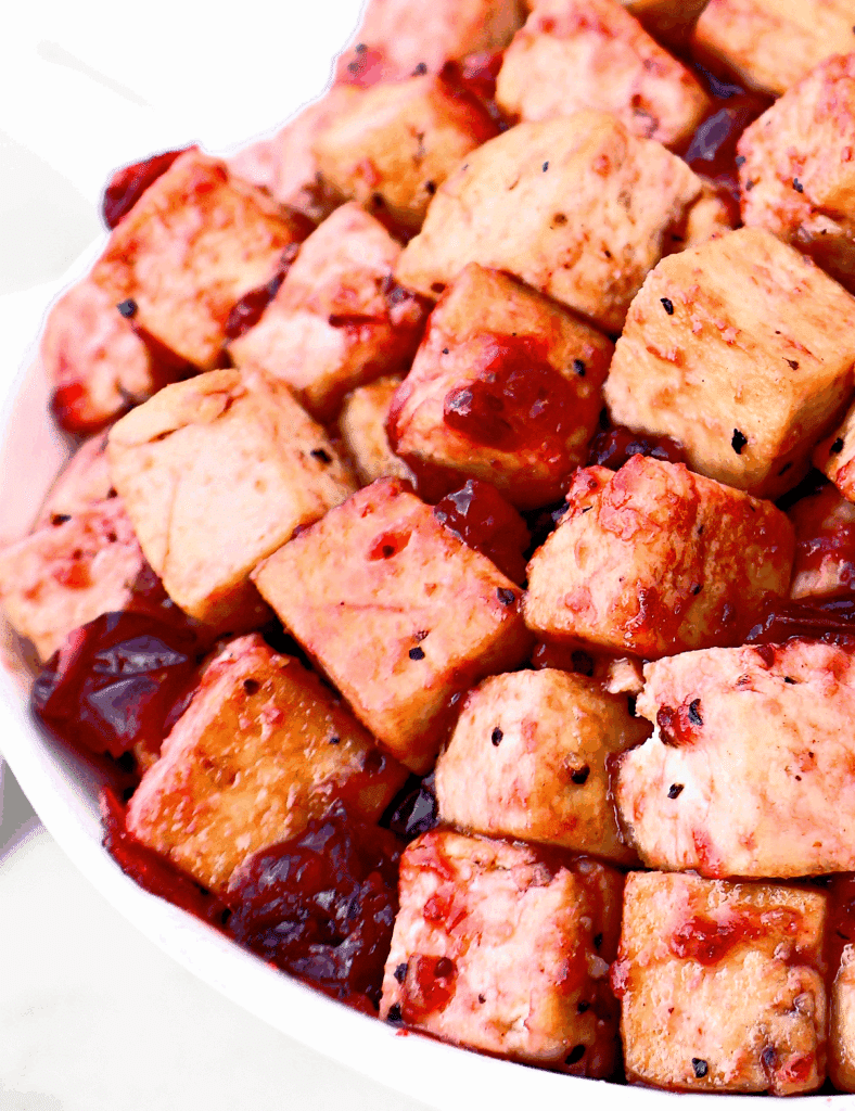 Close-up of golden-brown tofu cubes coated in a glossy red sauce, served in a white bowl. Some sauce pools around the tofu, giving a vibrant and appetizing appearance.