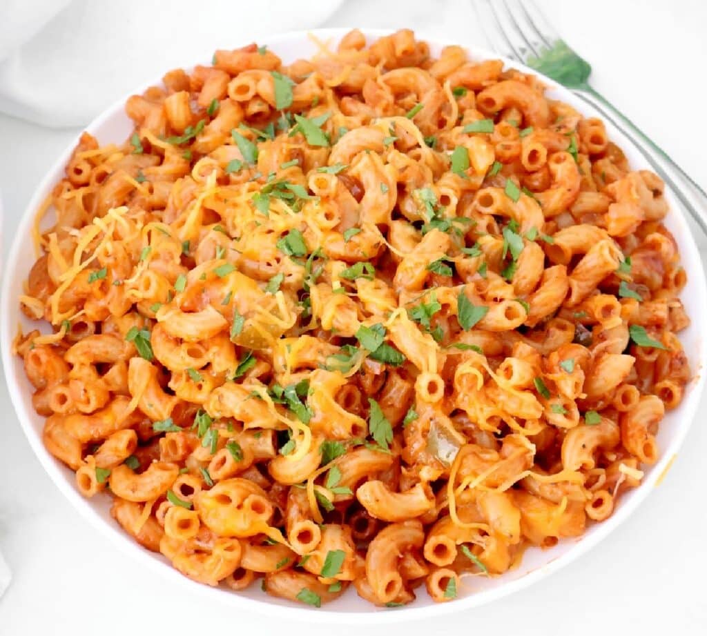 A white bowl filled with American goulash—macaroni noodles in rich red tomato sauce, topped with shredded cheese and chopped fresh herbs, with a fork to the side.