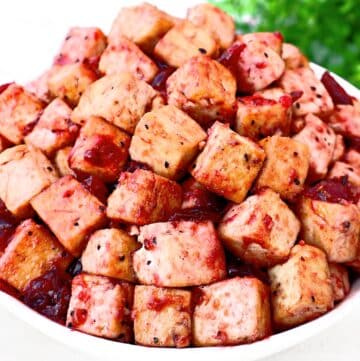 A bowl filled with cubed tofu coated in a reddish sauce, garnished with black pepper, with fresh green herbs blurred in the background.