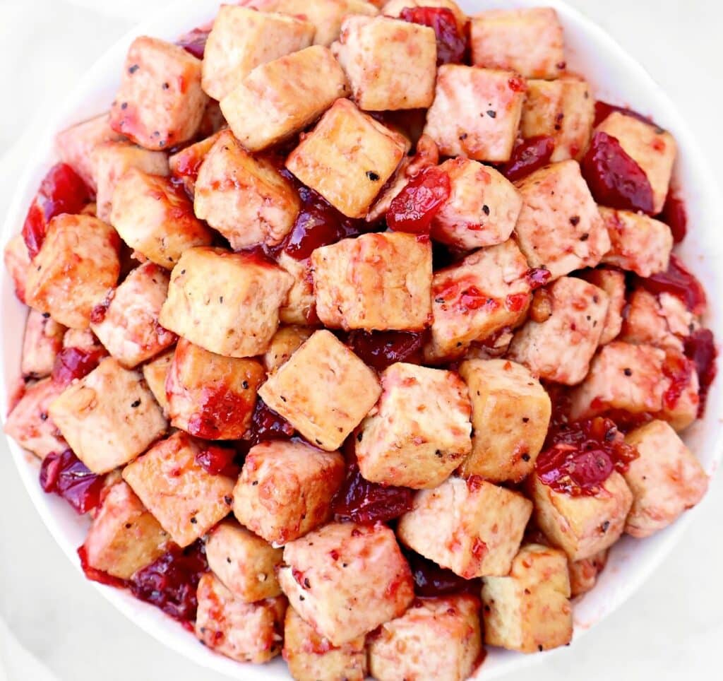A bowl filled with golden-brown tofu cubes coated in a red, glossy sauce with pieces of what appears to be dried cranberries or fruit.