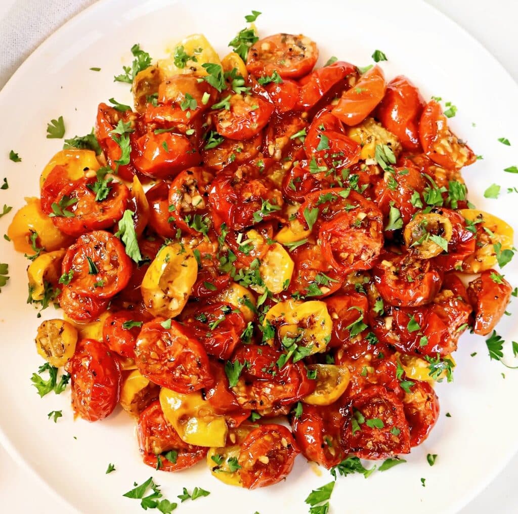 A white plate filled with roasted red and yellow cherry tomatoes, garnished with chopped fresh parsley.