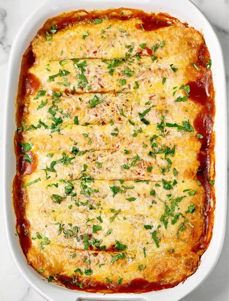 A baked casserole dish filled with enchiladas topped with melted cheese, chopped parsley, and red sauce, viewed from above on a white background.