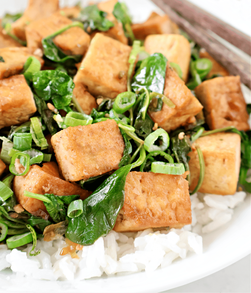 A close-up of a dish featuring golden-brown tofu cubes, sautéed spinach, and chopped green onions served over white rice, with chopsticks resting on the side.
