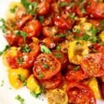 A close-up of roasted red and yellow cherry tomatoes garnished with fresh chopped parsley, served on a white plate.