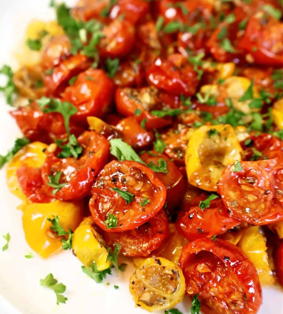 A close-up of roasted red and yellow cherry tomatoes garnished with fresh chopped parsley, served on a white plate.