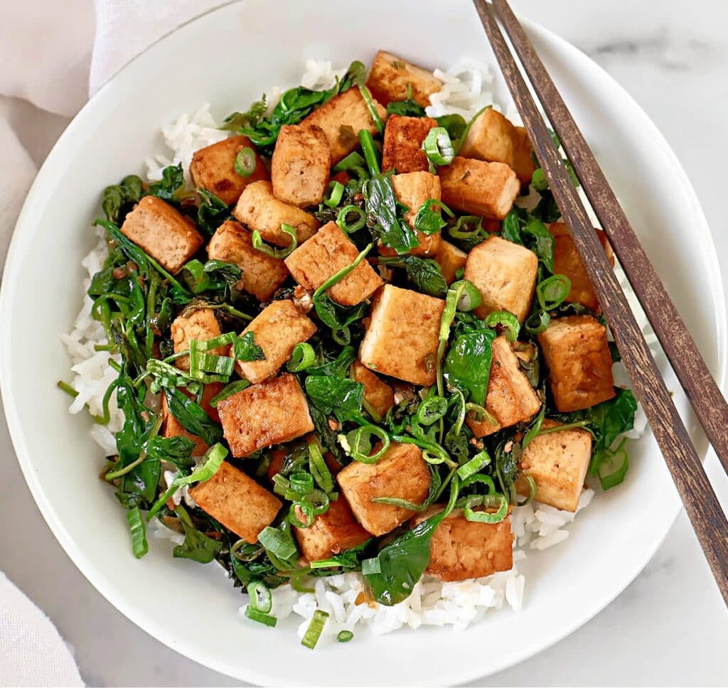 A white bowl filled with white rice, sautéed tofu cubes, leafy greens, and chopped green onions, with a pair of chopsticks resting on the rim.