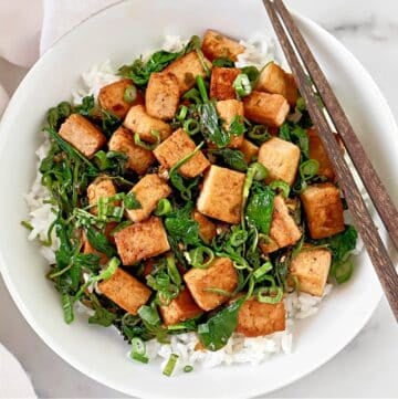 A white bowl filled with white rice, sautéed tofu cubes, leafy greens, and chopped green onions, with a pair of chopsticks resting on the rim.