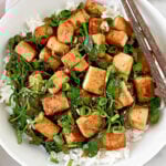 A bowl of white rice topped with sautéed tofu cubes, wilted spinach, and chopped green onions, with a pair of chopsticks resting on the side of the bowl.