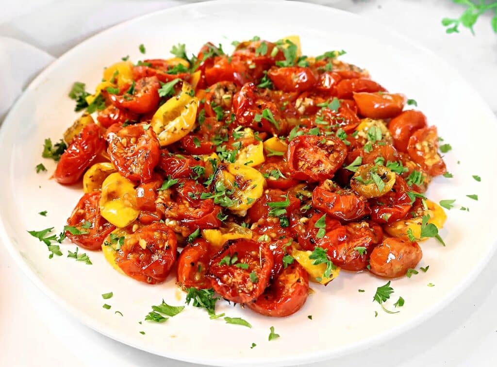 A white plate filled with roasted red and yellow cherry tomatoes, garnished with chopped fresh parsley and herbs. The tomatoes appear juicy and slightly caramelized.