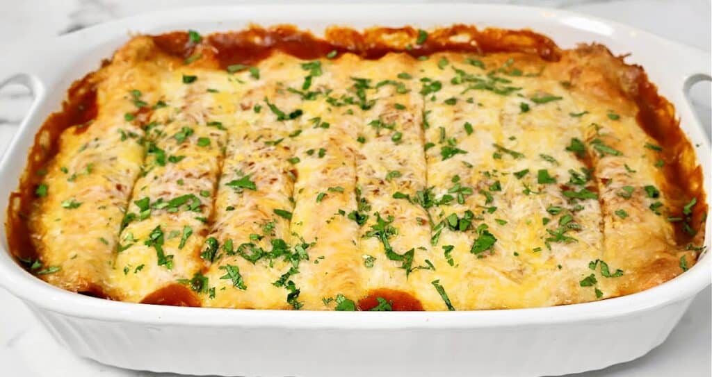 A white casserole dish filled with baked enchiladas, topped with melted cheese, red sauce, and chopped fresh cilantro.