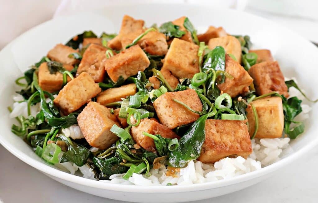 A white bowl filled with steamed white rice, topped with sautéed tofu cubes, wilted greens, and sliced green onions.