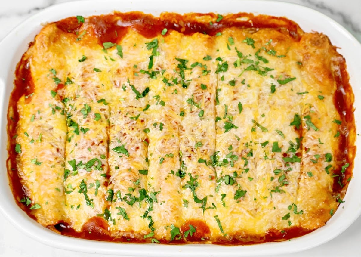 A baked dish of enchiladas topped with melted cheese and chopped cilantro, arranged in a white casserole dish with visible red sauce around the edges.