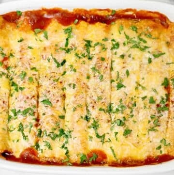 A baked dish of enchiladas topped with melted cheese and chopped cilantro, arranged in a white casserole dish with visible red sauce around the edges.