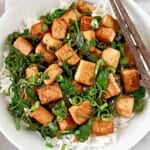 A bowl of white rice topped with sautéed tofu cubes, wilted spinach, and chopped green onions, with a pair of chopsticks resting on the side of the bowl.