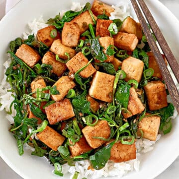 A bowl of white rice topped with sautéed tofu cubes, wilted spinach, and chopped green onions, with a pair of chopsticks resting on the side of the bowl.