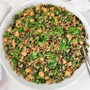 A white bowl filled with a colorful salad of quinoa, spinach, chickpeas, and herbs, with a serving spoon on the side.