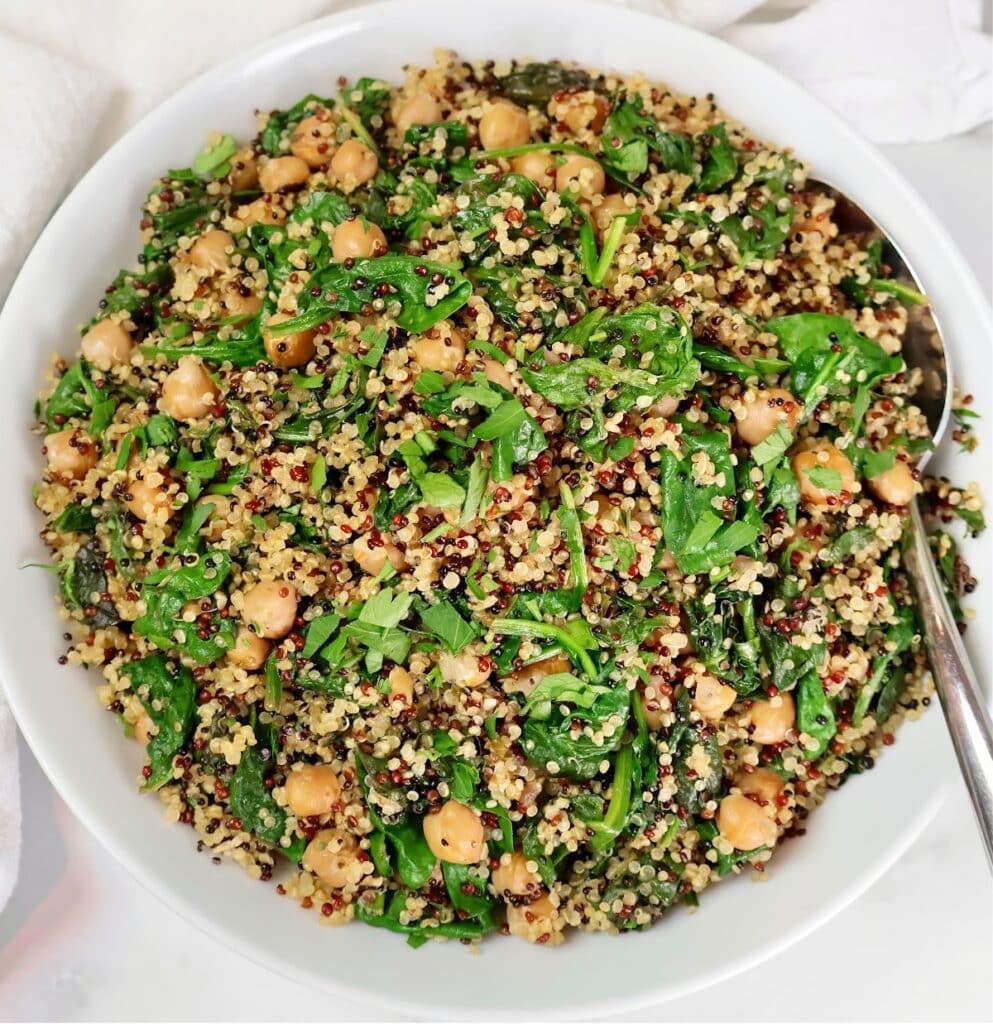 A white bowl filled with a colorful salad of quinoa, spinach, chickpeas, and herbs, with a serving spoon on the side.