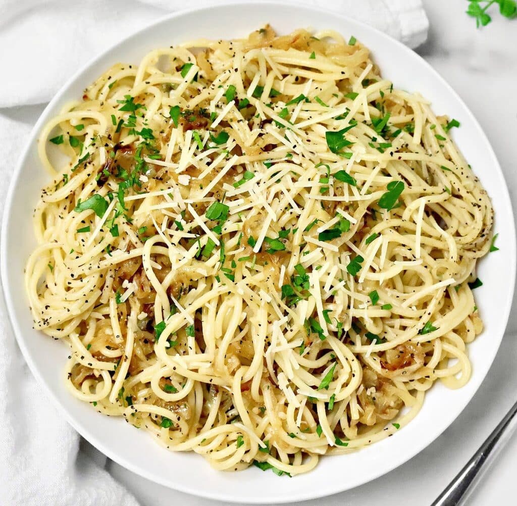A plate of spaghetti topped with grated cheese, chopped parsley, black pepper, and caramelized onions, served on a white dish with a fork nearby.