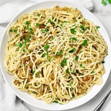 A plate of spaghetti topped with grated cheese, chopped parsley, black pepper, and caramelized onions, served on a white dish with a fork nearby.