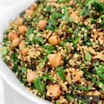 A close-up of a white bowl filled with quinoa, chickpeas, and fresh spinach, garnished with herbs. The dish appears colorful and healthy, showcasing green leaves and a mix of light and dark quinoa grains.