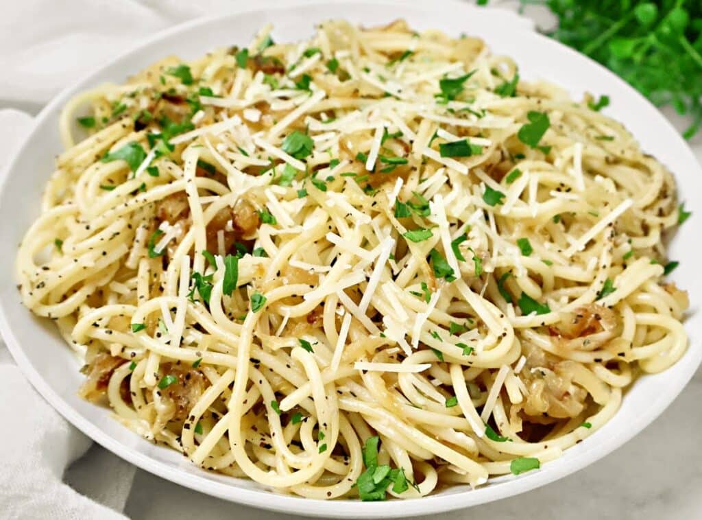 A plate of spaghetti topped with grated cheese, chopped parsley, caramelized onions, and ground black pepper, served on a white dish.