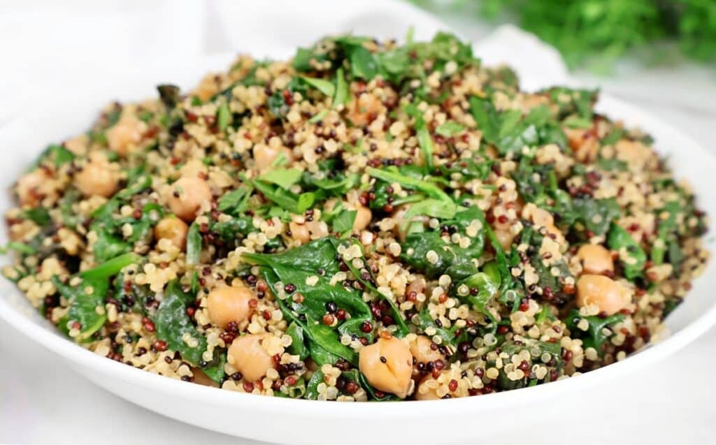 A white bowl filled with a salad made of quinoa, chickpeas, spinach, and fresh herbs, with a mix of light and dark quinoa grains visible.