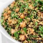 A close-up of a white bowl filled with quinoa, chickpeas, and fresh spinach, garnished with herbs. The dish appears colorful and healthy, showcasing green leaves and a mix of light and dark quinoa grains.