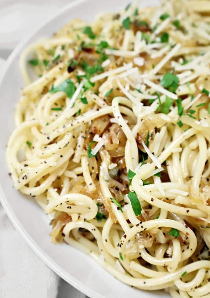 A close-up of a plate of spaghetti topped with grated cheese, chopped parsley, and cracked black pepper, with visible caramelized onions mixed into the pasta.