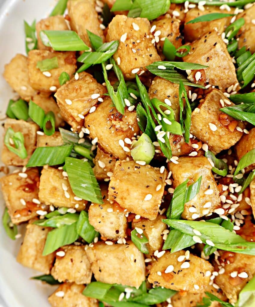 A close-up of crispy tofu cubes garnished with sliced green onions and sprinkled with sesame seeds on a white plate.
