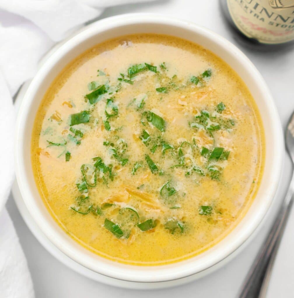 A bowl of creamy Guinness beer cheese soup garnished with chopped green herbs, placed on a white surface next to a spoon and a bottle.