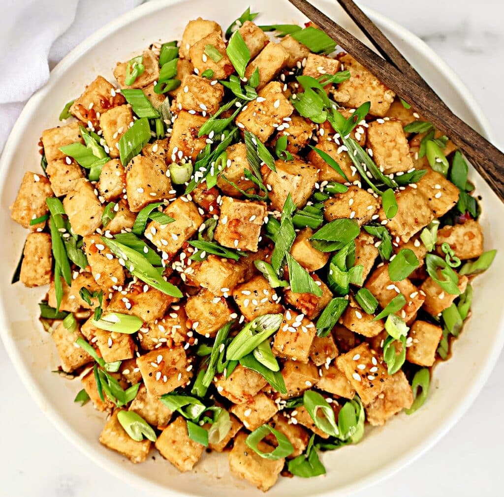 A bowl of crispy tofu cubes topped with sliced green onions and sesame seeds, with a pair of wooden chopsticks resting on the rim.