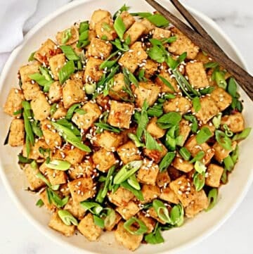 A bowl of crispy tofu cubes topped with sliced green onions and sesame seeds, with a pair of wooden chopsticks resting on the rim.