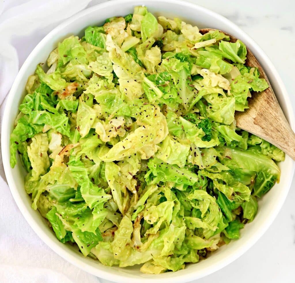 A white bowl filled with cooked, chopped cabbage seasoned with black pepper. A wooden spoon rests inside the bowl on the right side. The background is a light-colored surface.