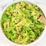 A white bowl filled with cooked, chopped cabbage seasoned with black pepper. A wooden spoon rests inside the bowl on the right side. The background is a light-colored surface.