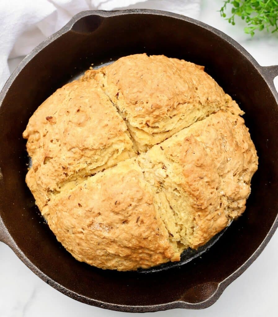 A round loaf of golden-brown Irish soda bread with caraway, featuring a cross cut into the top, baked in a black cast iron skillet on a white background.