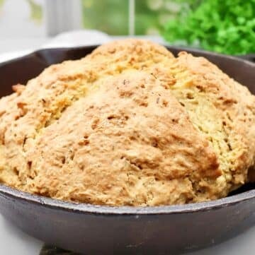 A round, golden-brown loaf of Irish soda bread with caraway sits in a black cast iron skillet, its textured, rustic crust inviting. Green leafy plants and a window are softly blurred in the background.