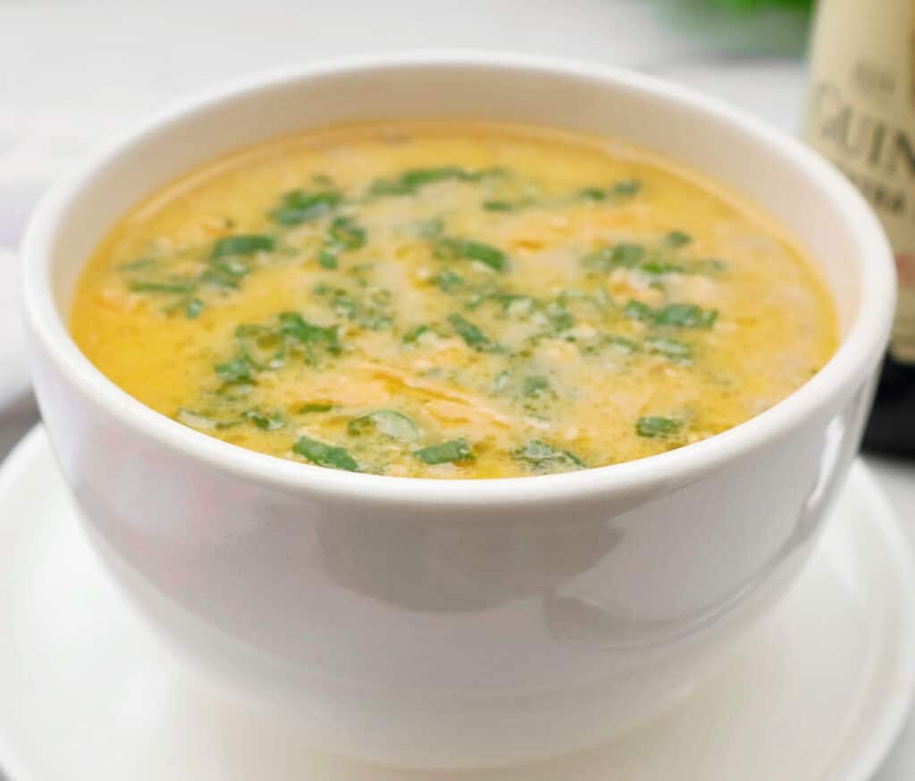 A white bowl filled with creamy yellow guinness beer cheese soup, garnished with chopped green herbs, placed on a matching saucer.