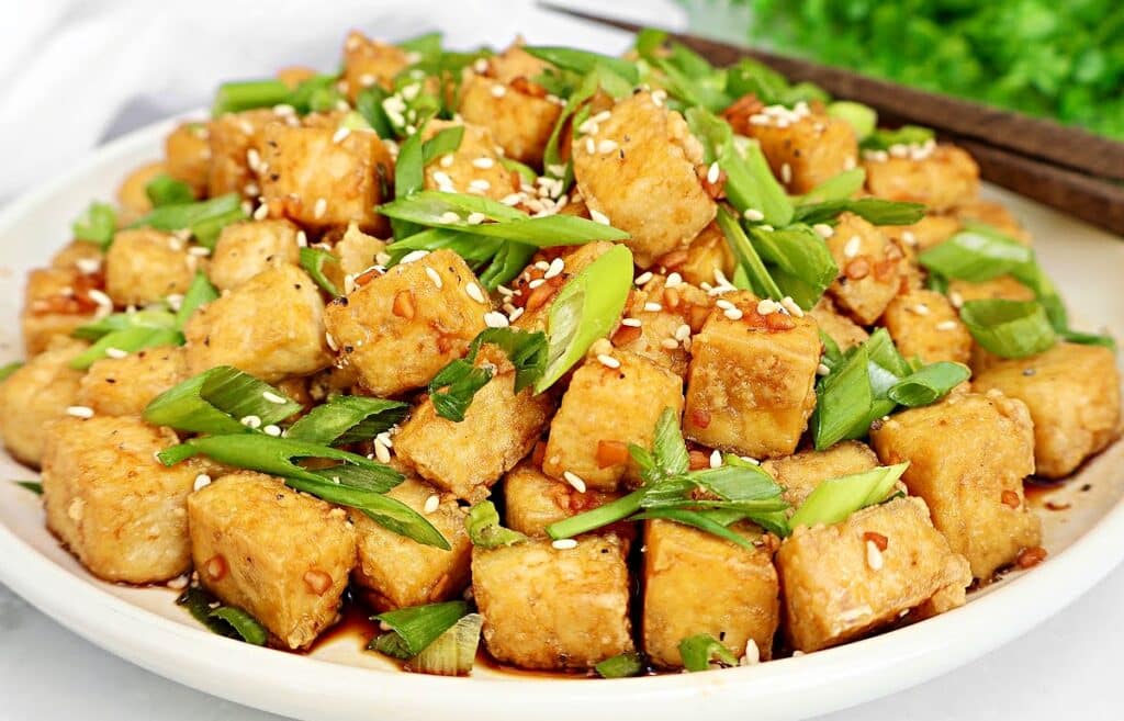 A plate of crispy golden tofu cubes topped with chopped green onions, sesame seeds, and a drizzle of sauce, served on a white dish.