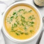 A bowl of creamy Guinness beer cheese soup garnished with chopped green herbs, placed on a white surface next to a spoon and a bottle.