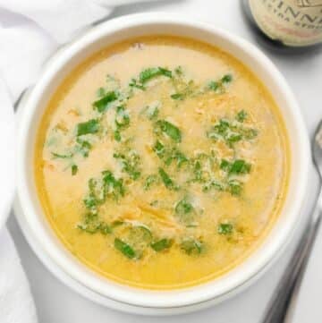 A bowl of creamy Guinness beer cheese soup garnished with chopped green herbs, placed on a white surface next to a spoon and a bottle.
