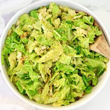A white bowl filled with cooked, chopped cabbage seasoned with black pepper. A wooden spoon rests inside the bowl on the right side. The background is a light-colored surface.