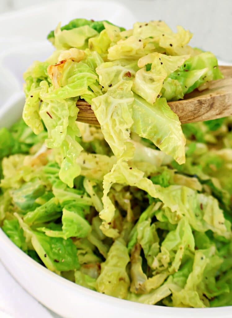 A wooden spoon lifts a serving of cooked, seasoned cabbage from a white bowl filled with shredded green cabbage. The cabbage appears tender and lightly coated with spices.